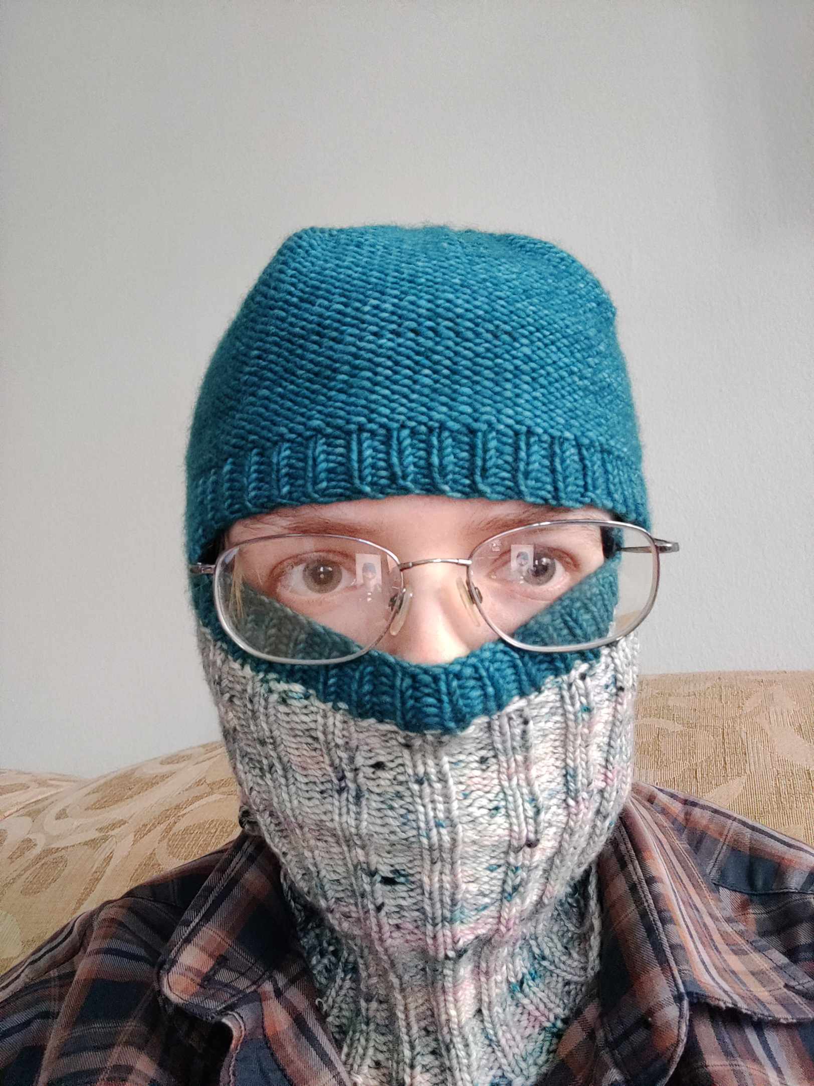 Emma wearing a knit balaclava. The balaclava is turquoise on the top and variegated white with bits of turquoise and pink on the bottom. The neck and lower face portion is a 2 by 2 rib, the eye hole is surrounded by a 1 by 1 rib, and the crown of the hat is stockinette.