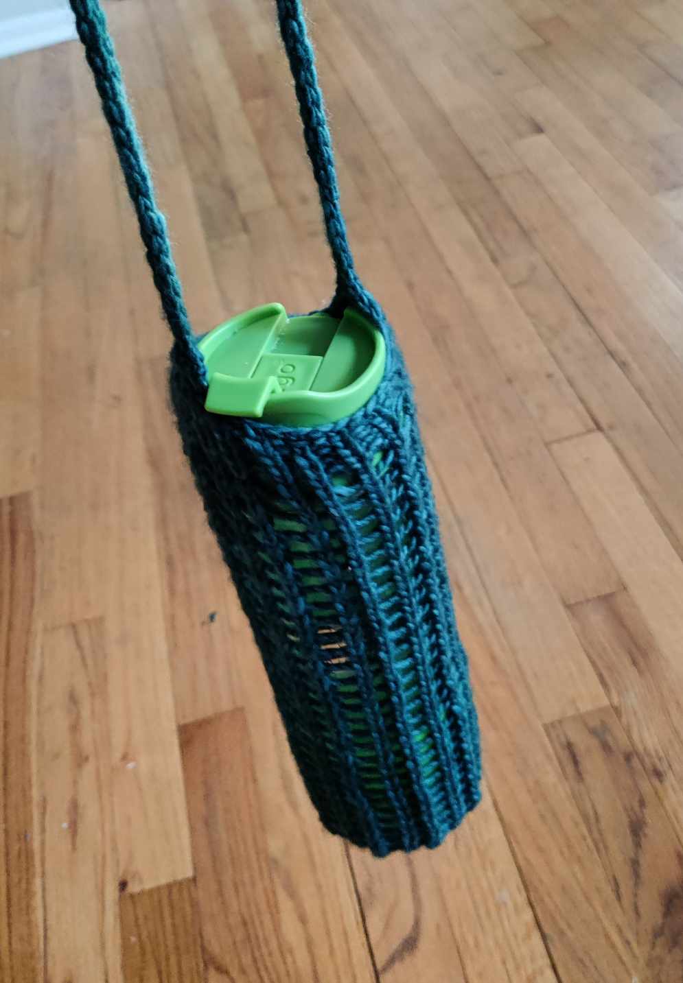 A water bottle held in a knit mesh sleeve with a grid-like lace pattern and an icord shoulder strap, knit in the same turquoise yarn as the balaclava