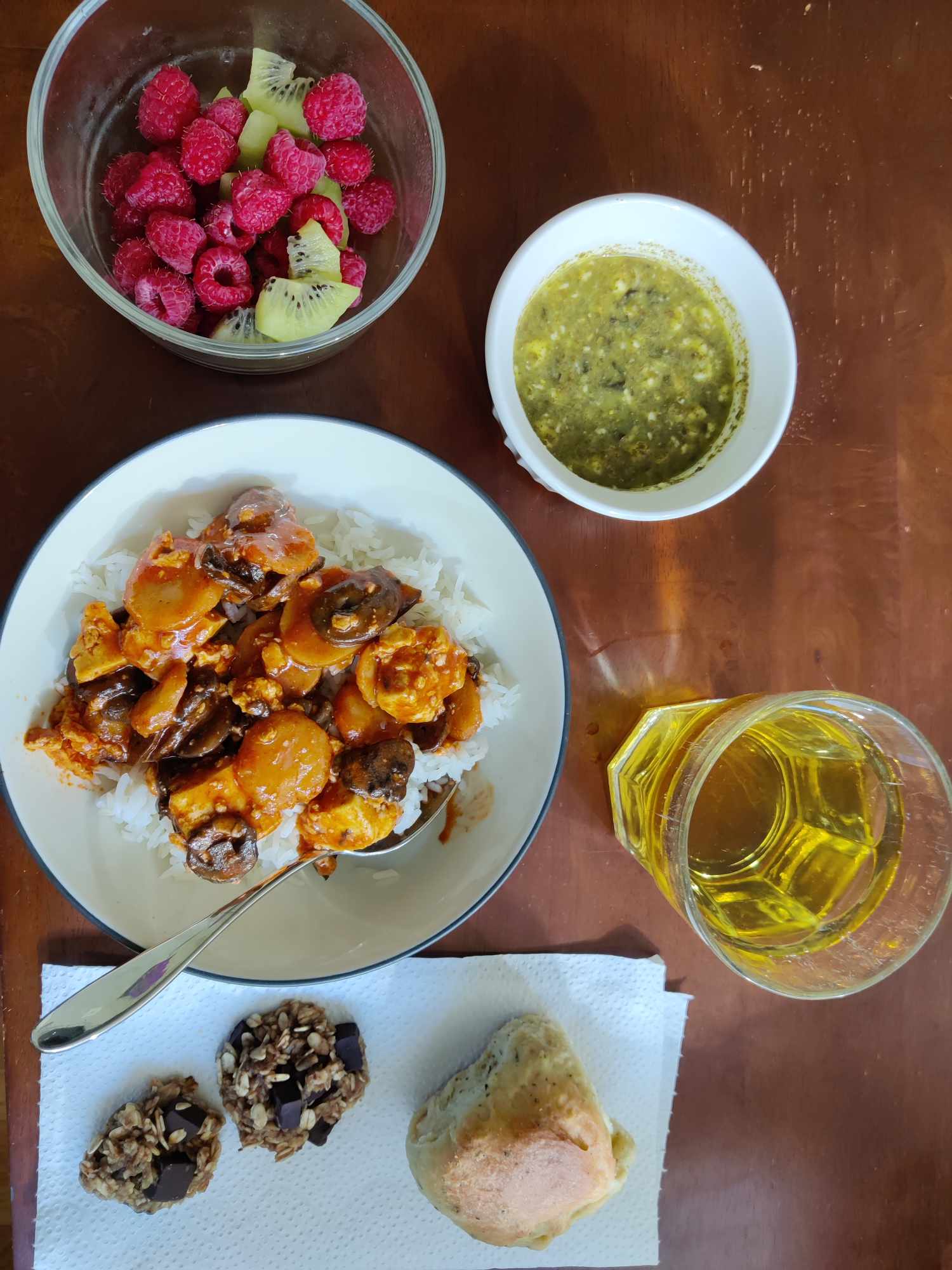 A fruit salad including raspberries and kiwis,
a small bowl of pesto dip, a bowl of white rice with tofu and vegetables in an orange sauce,
a glass of apple juice, a bread roll, and two small chocolate chip oat cookies