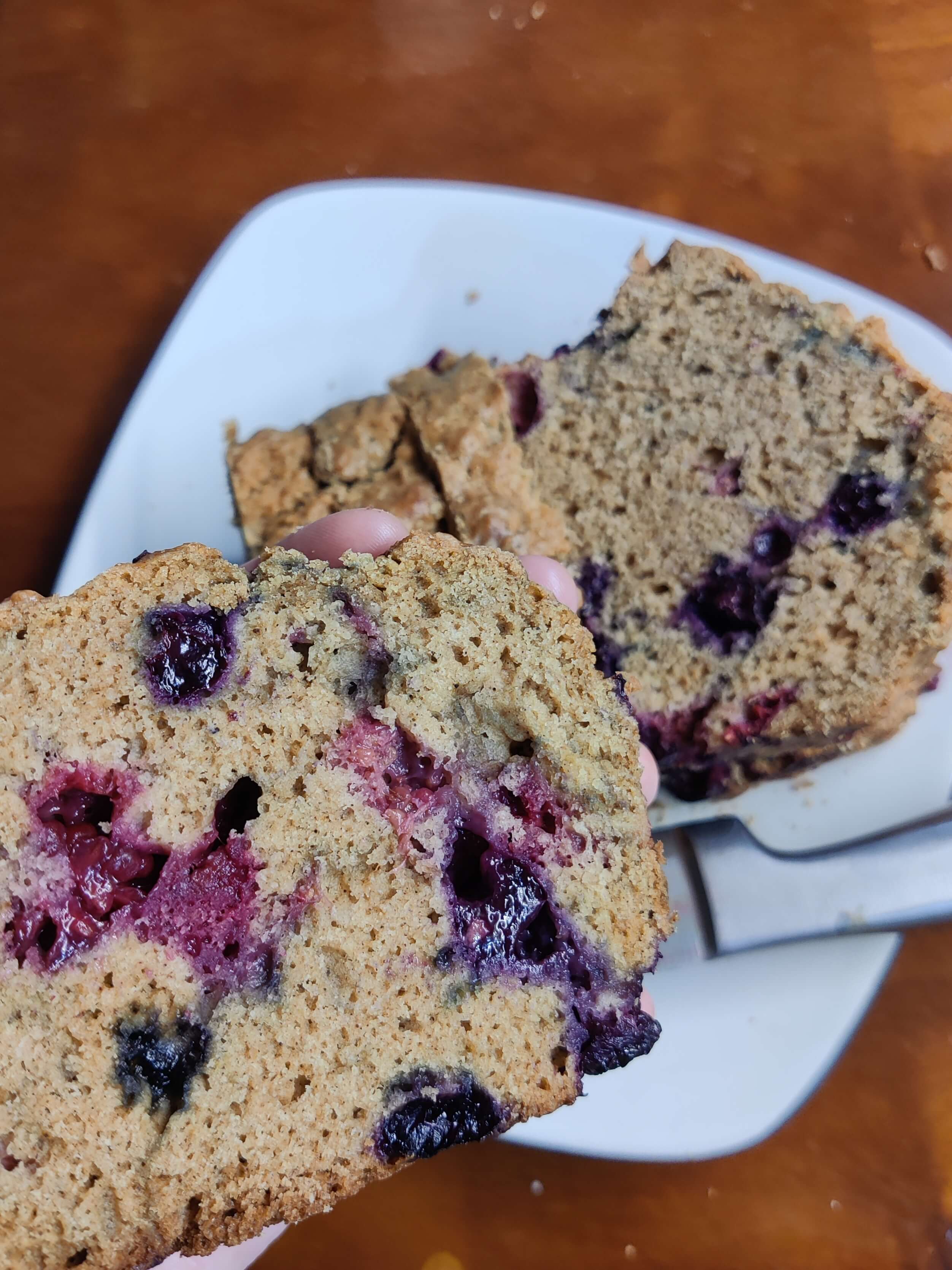 A hand holds a slice of bread up to the camera. The bread is light brown and has berries in it spotting the bread with bluish/pinkish purple. In the background you can see the full sliced loaf on the plate.